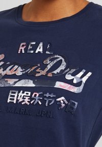 Námornícky modré bavlnené tričko s vyvýšeným kvetinovým potlačou písmen "REAL SuperDry" a ďalším textom v rôznych textúrach a farbách.
