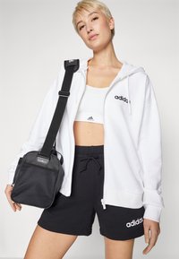 Fehér cipzáros kapucnis felső fekete adidas logóval, fekete sorttal és hálós részletekkel ellátott kisméretű fekete táskával, sportos fehér toppal viselve.