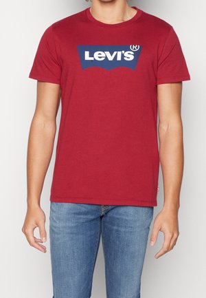 Person wearing a red Levi’s t-shirt with a blue and white logo, paired with blue jeans, standing against a plain background.