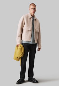 Veste en polaire beige texturée avec un col et deux poches avant, associée à une chemise bleu clair et un jean noir, tenant un pull jaune.