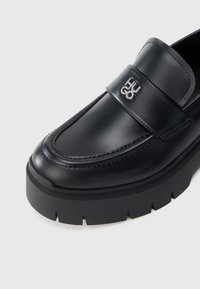 Musta nahkainen loaferi paksulla pohjalla ja hopealla HU 03 -logolla nauhassa yläpuolella.