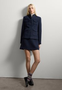 Abito in tweed blu navy con giacca aderente dotata di quattro tasche e gonna dritta. Il modello indossa calzini a righe in maglia e mocassini neri.