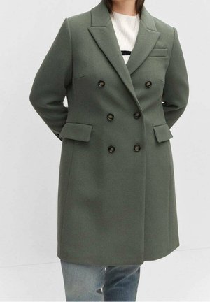 Femme portant un manteau vert olive à double boutonnage avec de larges revers, par-dessus un pull blanc et un jean bleu, mains derrière le dos.