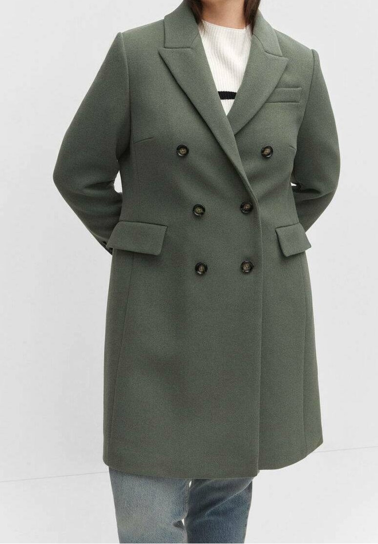 Femme portant un manteau vert olive à double boutonnage avec de larges revers, par-dessus un pull blanc et un jean bleu, mains derrière le dos.