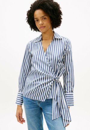 Frau trägt eine blau-weiß gestreifte Wickelbluse mit vertikalen Streifen, die an der Taille zu einer Schleife gebunden ist, kombiniert mit hellblauen Jeans, steht vor weißem Hintergrund.