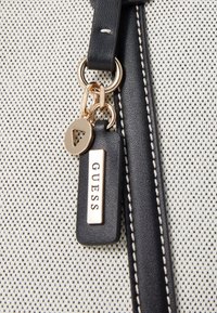 Sac à main en tissu à motifs noir et blanc avec un porte-clés en accent doré, comportant une étiquette rectangulaire avec "GUESS" en lettres dorées.