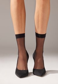 Svarta högklackade skor med spetsig tå, kombinerade med genomskinliga svarta ankelsockor med en solid resårband upptill. Slät yta, klassisk design.