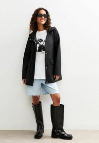 Czarna wodoodporna kurtka z kapturem, biała koszulka z grafiką, jasne dżinsowe szorty i czarne kalosze do kolan z dekoracyjnymi klamrami.