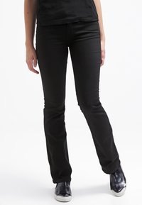 Personne portant un pantalon noir ajusté, un haut noir et des baskets noires à semelles blanches, se tenant sur un fond blanc.