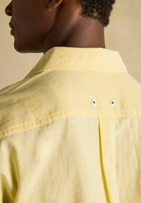 Osoba ubrana w jasnżółtą lnianą koszulę z dwoma małymi białymi otworami wentylacyjnymi na górnej części pleców, zbliżenie od tyłu.