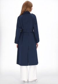 Marineblauwe trenchcoat met een riem, lange mouwen en een plooi aan de achterkant. Gladde stof met een klassiek ontwerp, gecombineerd met witte broek met wijde pijpen.