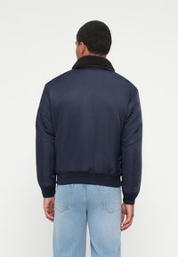 Veste bomber bleu marine avec un col en polaire marron, des poignets et une taille côtelés ; texture lisse, design minimaliste et coupe ajustée.