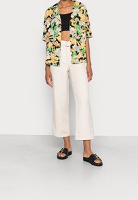 Kwiatowa koszula z krótkim rękawem w kolorze czarnym, zielonym, pomarańczowym i kremowym; czarny top krótki; kremowe spodnie typu wide-leg; czarne sandały z złotą klamrą.