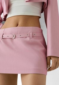 Mini gonna rosa con una texture liscia, dotata di una cintura larga con hardware argentato e passanti per la cintura. Indossata con un top corto bianco.
