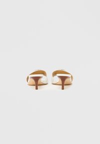 Bijele papuče s potpeticom, otvorenim stražnjim dijelom i zaobljenim prstima. Svijetla drvena potpetica s rebrastom teksturom koja kontrastira s glatkom površinom cipela.