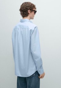 Jasnoniebieska koszula z długim rękawem, luźny krój, z plisą z tyłu, w połączeniu z ciemnoniebieskimi dżinsami i okularami w kolorze tortoise.