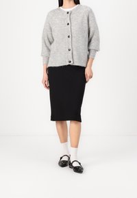 Ljusgrå stickad cardigan med rund hals, sex mörka knappar, bärs över en svart knälång kjol, tillsammans med vita strumpor och svarta skor.
