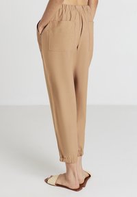 Pantaloni beige tapered con cintura elastica e polsini, dotati di due tasche posteriori e una texture liscia, abbinati a sandali scivolati chiari.