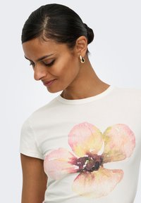 Femme aux cheveux foncés en chignon, portant des boucles d’oreilles en or et une chemise blanche avec un grand motif floral aquarelle rose et jaune.