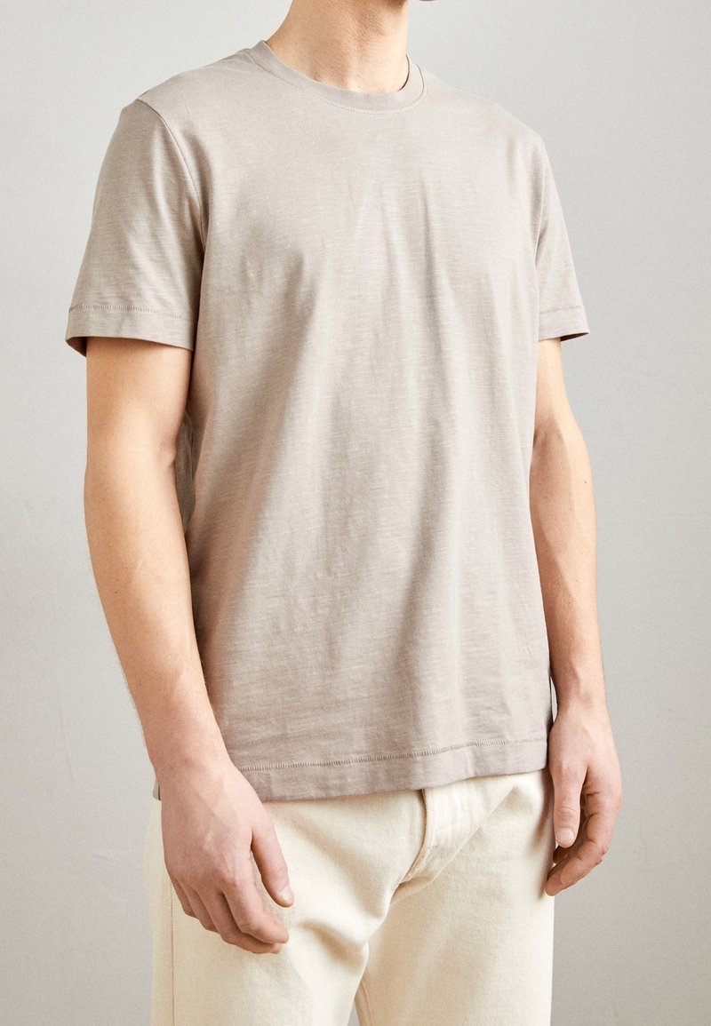 Homme portant un t-shirt à manches courtes beige clair et un pantalon blanc cassé, se tenant contre un fond clair et uni.