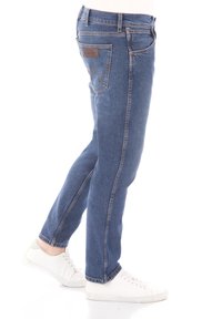 Blauwe denim jeans met een slim fit, voorzien van contrasterende stiksels en een klein leren label op de achterzak. Draagt met witte sneakers.