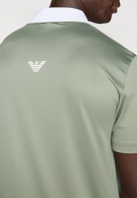 Πόλο μπλούζα σε απαλό πράσινο ύφασμα, με λευκό γιακά και μικρό λευκό λογότυπο στην πλάτη. Κοντά μανίκια, υφή υλικού, άνετη εφαρμογή.