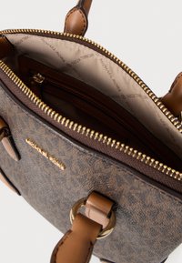 Καφέ τσάντα χειρός με σχέδιο, χρυσά μεταλλικά στοιχεία, κλείσιμο με φερμουάρ και μπεζ λουράκι. Εσωτερικό με φόδρα που διαθέτει τσέπη με φερμουάρ και λεπτομέρειες με λογότυπο.