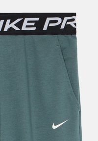 Grønne Nike Pro joggebukser med svart midjebånd som har stor hvit "NIKE PRO" tekst og hvit Nike swoosh-logo nær lommen.