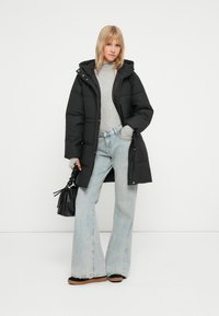 Čierna prešívaná bunda s kapucňou, so zipsom a vreckami, kombinovaná s svetlomodrými širokými nohavicami a malou čiernou kabelkou.