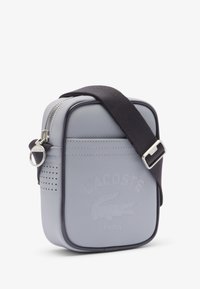 Borsa a tracolla Lacoste grigio chiaro con superficie testurizzata, zip argento, tracolla nera e logo in rilievo sul fronte. Forma quadrata.