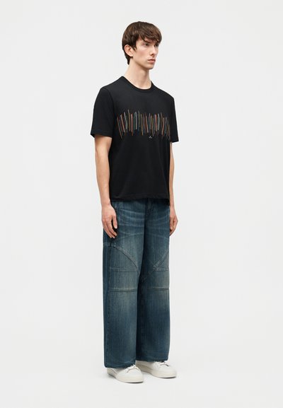 Νέος άνδρας στέκεται φορώντας μαύρο μπλουζάκι με σχέδιο, πλατύτερο τζιν σκουρομπλε με ραφές, και λευκά αθλητικά, μπροστά σε απλό φόντο.