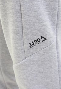 Tissu gris clair avec une étiquette imprimée noire "JJ90" et les coordonnées 55.9847°N 9.1288°E sur un détail de couture.