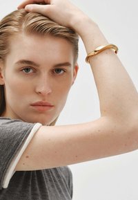 Brazalete de metal dorado en la muñeca de un modelo. El brazalete presenta un diseño liso y ondulado. El modelo lleva una camiseta gris con detalles blancos.