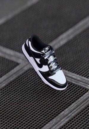 Czarno-biała tenisówka Nike z szarymi sznurowadłami i czarnym zawieszonym uśmiechem, spoczywająca na metalowej kratce podłogowej.