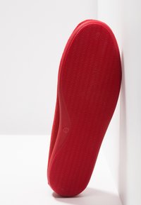 Chausson en suède rouge avec une semelle en caoutchouc texturé ; présente des bords lisses et arrondis ainsi qu'un motif ondulé discret sur le dessous pour une meilleure adhérence.