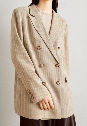 Person iført en beige dobbeltradet blazer med brune knapper og et skjorte i neutrale toner under, mod en ensfarvet baggrund.