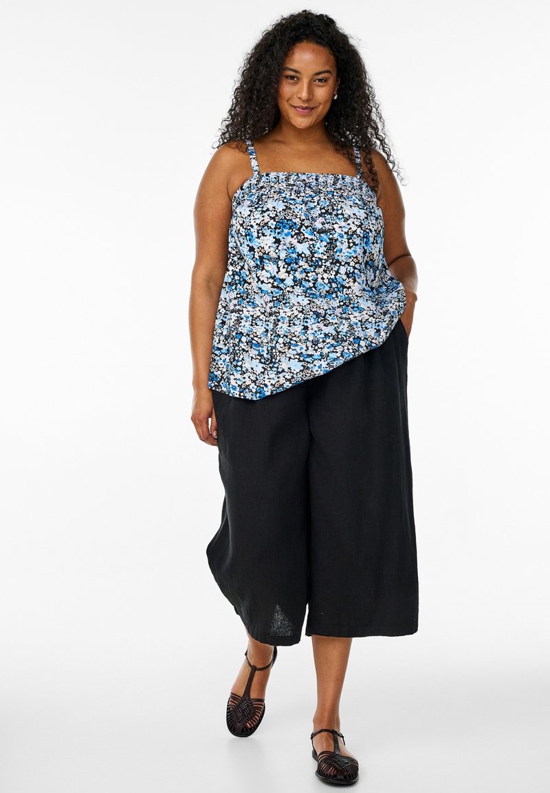 Donna con i capelli ricci che indossa un top smanicato blu con motivo floreale, pantaloni neri a gamba larga tagliati e scarpe nere piatte con cinturini, che cammina in avanti.