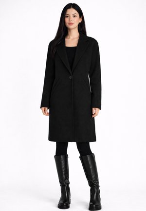 Donna che indossa un lungo cappotto nero monopetto con rever a lancia, leggings neri e stivali neri in pelle fino al ginocchio, in piedi su sfondo bianco.