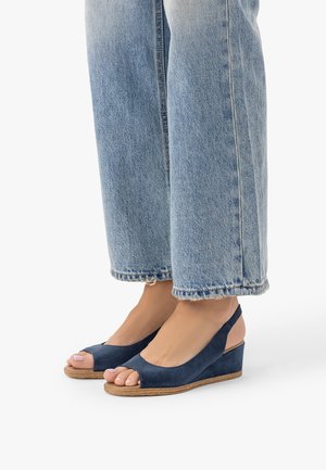 Inimene, kes kannab helehalli lühendatud teksapükse ja tumesiniseid avatud varbaga kiilkontsaga sandaale rihmadega, seisab valgel pinnal.