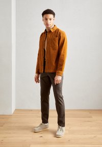 Brown corduroy shirt jacket with a chest pocket, paired with dark trousers and light gray shoes, on a wooden floor background.