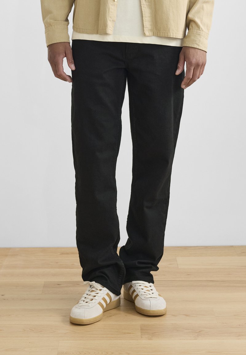 Sorte rette bukser laget av denim, parret med hvite sneakers med gullstriper. En lys beige kordfløyelskjorte er iført over en kremfarget skjorte.