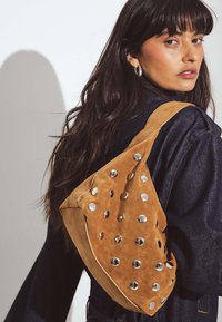 Borsa in suede marrone con una forma unica, caratterizzata da accenti circolari in argento e una tracolla ampia. La superficie texture aggiunge dettaglio e interesse visivo.