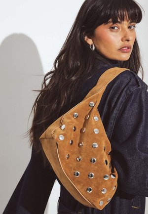Pruun suede kott unikaalse kuju ja hõbedaste ümmarguste detailidega ning laia rihmaga. Tekstureeritud pind lisab detaili ja visuaalset huvi.