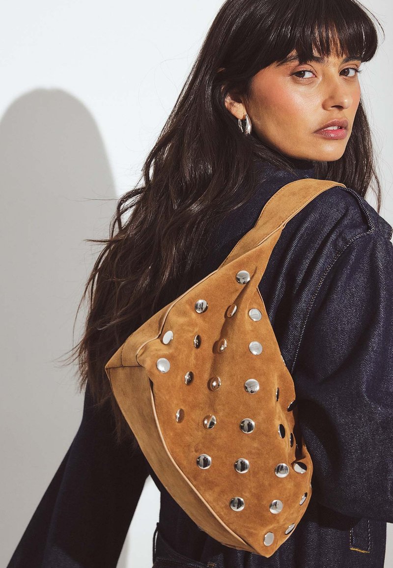 Borsa in suede marrone con una forma unica, caratterizzata da accenti circolari in argento e una tracolla ampia. La superficie texture aggiunge dettaglio e interesse visivo.