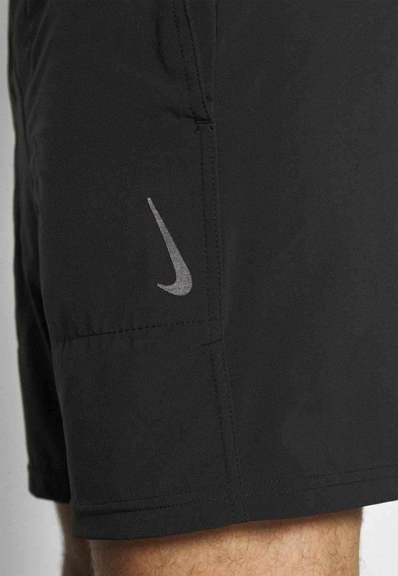Shorts de sport noirs avec le logo Nike en forme de virgule gris sur la cuisse gauche, portés par une personne avec des poils visibles sur le bas de la jambe.