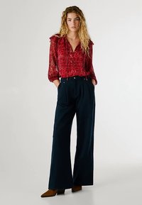 Rdeča bluza z vzorcem in nabranimi rokavi nad temnimi širokimi hlačami. Model nosi rjave čevlje s peto. Gladek material in sproščen kroj.