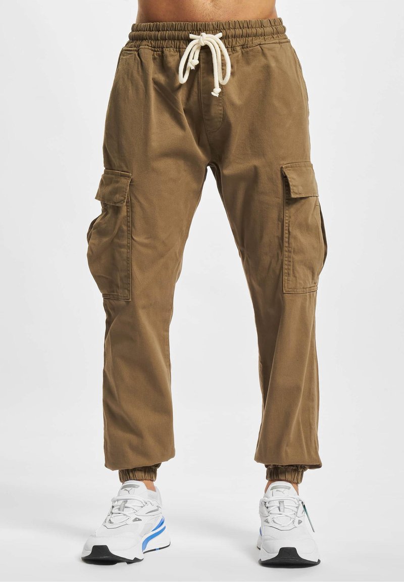 Pantalon cargo marron avec taille et poignets élastiques, poches avant à rabat, et baskets blanches avec détails bleus sur une personne debout.