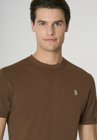 Maglione lavorato a maglia marrone a maniche corte con scollatura a costine e dettaglio logo discreto sul petto. Texture liscia, design aderente.