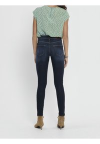 Femme portant un jean skinny bleu foncé, un chemisier à manches courtes vert clair avec des taches blanches, et des bottines en daim beige, vue de dos.