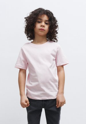 Lichtroze katoenen t-shirt met korte mouwen, ronde hals en een relaxte pasvorm, gecombineerd met donkergrijze broek. Model staat met de armen gekruist.
